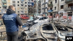 This photograph taken and released by Donetsk Region Prosecutor's Office on March 10, 2024 shows a war crimes prosecutor near destroyed cars in the courtyard of a damaged residential building following a missile attack in Myrnohrad, Donetsk region.