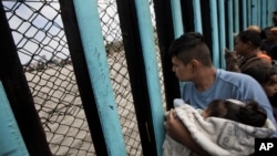 Pripadnik karavana migranata iz Centralne Amerike, držeći dijete, gleda kroz pograničnu ogradu prema grupi ljudi koji su se okupili na američkoj strani, dok stoji na plaži gde se pogranična žica završava u okeanu, u Tihuani, Meksiko, 29. aprila 2018.